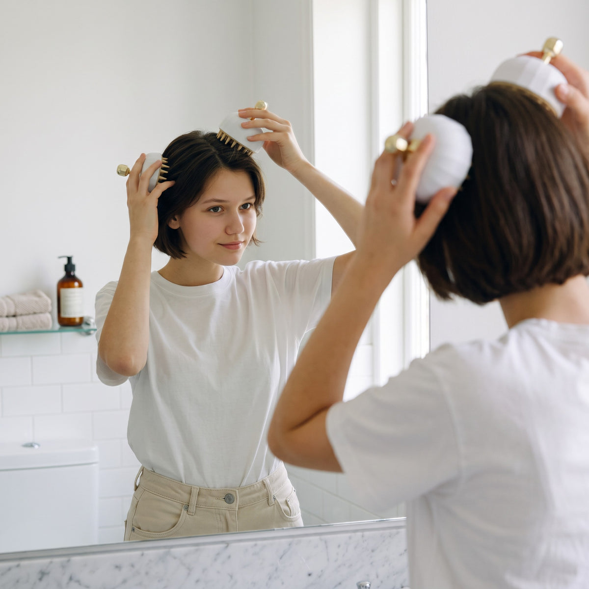 Mujer usando el cepillo masajeador zafiropulse Glow Brush blanco con detalles dorados frente al espejo del baño