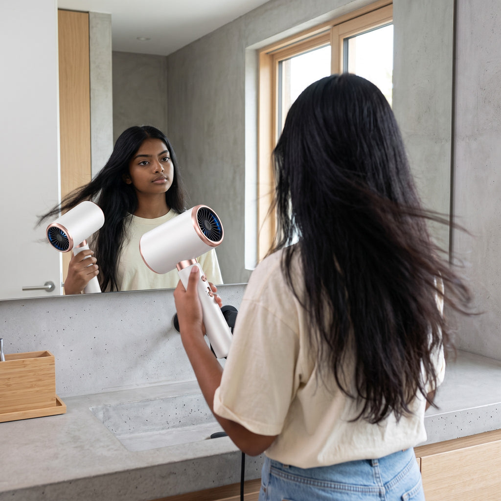 Mujer joven usando el secador de pelo zafiropulse PERENNE Aero-Glow Therapy blanco con detalles en oro rosa frente al espejo