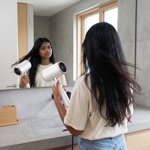 Mujer joven usando el secador de pelo zafiropulse PERENNE Aero-Glow Therapy blanco con detalles en oro rosa frente al espejo